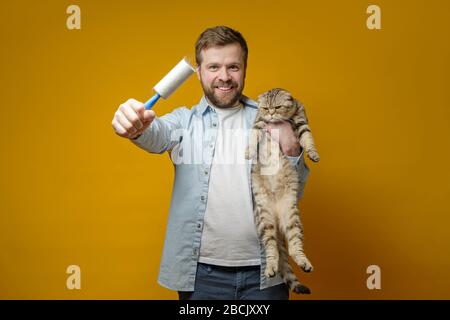 L'uomo contentato e sorridente tiene in mano un rullo con nastro adesivo nei capelli degli animali, nell'altro abbraccia il suo amato gatto. Foto Stock