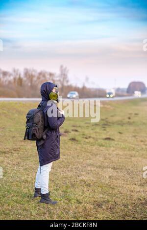 COVID-19. Una ragazza in abiti scuri, una maschera a gas e un cappuccio sulla testa. Giorno nuvoloso con la città. Ragazza viaggi hitchhiking. Fuga dal coronavirus i Foto Stock