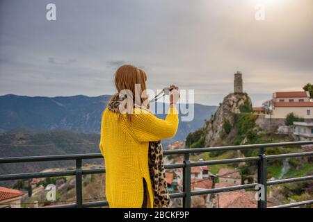 Giovane donna con un maglione giallo scatta foto di un pittoresco villaggio montano Foto Stock