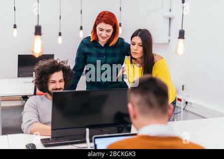 Due giovani donne d'affari accanto a un collega maschile mentre spiega le nuove attività online su un computer in un ambiente di ufficio confortevole. Foto Stock