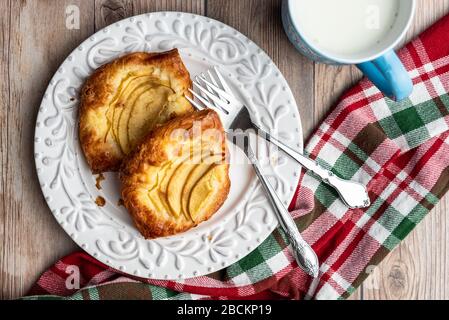 Crostate di mele singole fresche su un piatto bianco con forchette, tazza di latte, tovagliolo rosso, verde e bianco, sfondo di legno Foto Stock