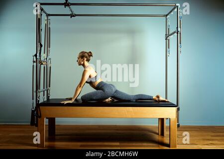 Giovane ragazza che fa esercizi pilates con un letto riformatore. Beautiful slim fitness trainer su sfondo grigio riformatore, tasto basso, luce arte. Fitness Foto Stock