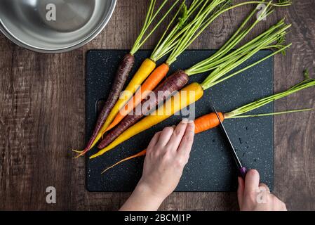 Mani donna taglio carote arcobaleno su tagliere nero, ciotola in acciaio inox, tavolo in legno Foto Stock