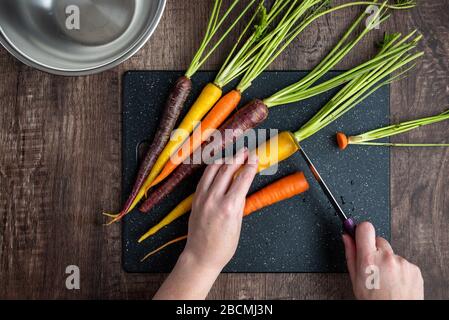 Mani donna taglio carote arcobaleno su tagliere nero, ciotola in acciaio inox, tavolo in legno Foto Stock