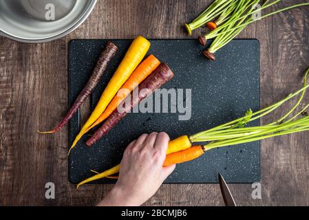 Mani donna taglio carote arcobaleno su tagliere nero, ciotola in acciaio inox, tavolo in legno Foto Stock