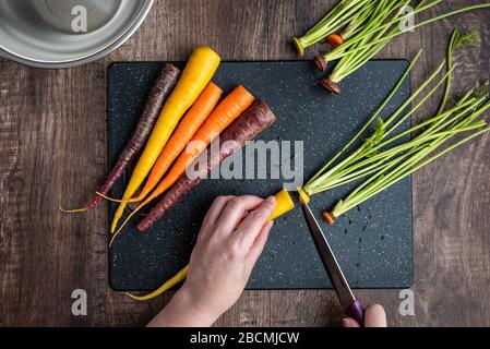 Mani donna taglio carote arcobaleno su tagliere nero, ciotola in acciaio inox, tavolo in legno Foto Stock