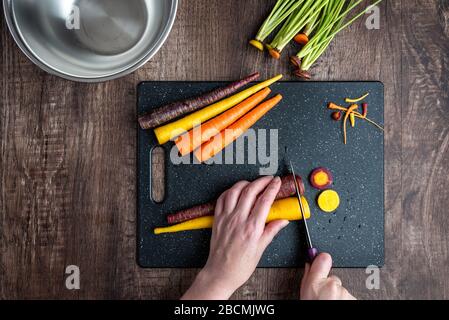 Mani donna taglio carote arcobaleno su tagliere nero, ciotola in acciaio inox, tavolo in legno Foto Stock