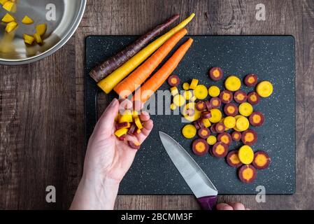 Mani donna taglio carote arcobaleno su tagliere nero, ciotola in acciaio inox, tavolo in legno Foto Stock