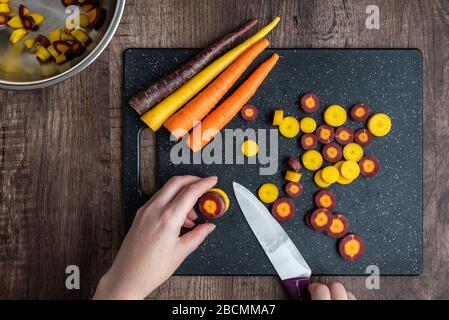 Mani donna taglio carote arcobaleno su tagliere nero, ciotola in acciaio inox, tavolo in legno Foto Stock