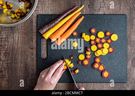 Mani donna taglio carote arcobaleno su tagliere nero, ciotola in acciaio inox, tavolo in legno Foto Stock