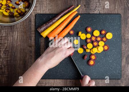 Mani donna taglio carote arcobaleno su tagliere nero, ciotola in acciaio inox, tavolo in legno Foto Stock