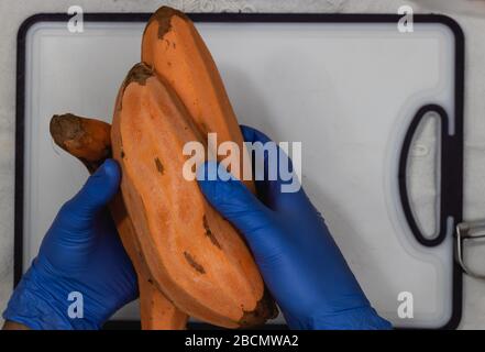 Un uomo con guanti blu tiene le patate dolci pelate, salvaguardie e cautela contro il virus corona all'interno della cucina domestica Foto Stock