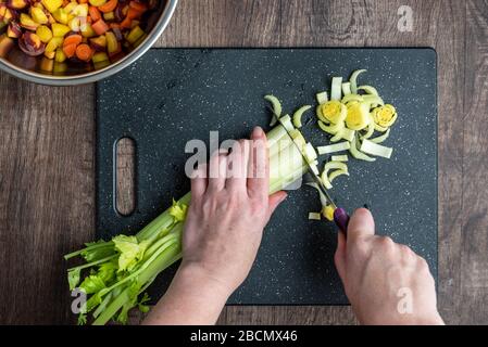 Le mani della donna tagliano un mazzo di sedano, tagliere nero e coltello da cuoco, ciotole di carote a dadini arcobaleno Foto Stock