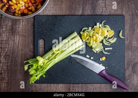Gambi di sedano, con qualche tritato, su un tagliere nero, coltello da cuoco, tavolo di legno, ciotola di carote a dadini arcobaleno Foto Stock