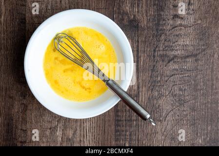 Uova sbattute crude in ciotola bianca con frusta di metallo, tavola di legno Foto Stock