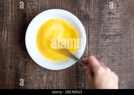 Mano da donna che monta uova crude in ciotola bianca con frusta di metallo, tavola di legno Foto Stock