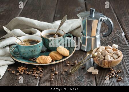 Due tazze vintage blu di caffè nero, biscotti e macchina da caffè geyser circondato da tovaglioli di lino grigio, pezzi di zucchero e chicchi di caffè su vecchia t di legno Foto Stock