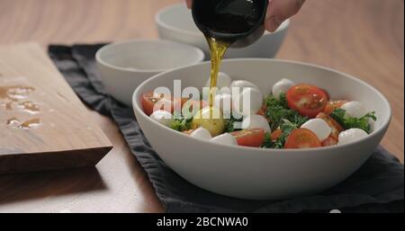 le mani dell'uomo versano il condimento sull'insalata con kale, pomodori ciliegini e mozzarella in una ciotola bianca Foto Stock