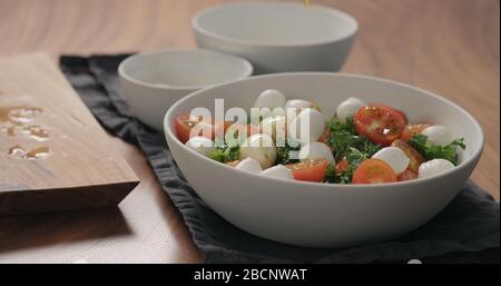 le mani dell'uomo versano il condimento sull'insalata con kale, pomodori ciliegini e mozzarella in una ciotola bianca Foto Stock