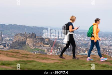 Edimburgo, Scozia, Regno Unito. 5 aprile 2020. La seconda domenica del blocco del coronavirus nel Regno Unito il pubblico è al di fuori di prendere il loro esercizio quotidiano. Donne che camminano su Salisbury Crags in Holyrood Park con il Castello di Edimburgo in lontananza. Iain Masterton/Alamy Live News Foto Stock