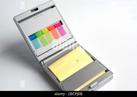 Distributore d'argento di note postit gialle e evidenziatori a indice appiccicoso multicolore su sfondo bianco, punto interrogativo scritto sulla nota postit. Foto Stock