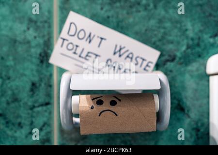 Rotolo di carta igienica rifinito in bagno con un'immagine di una faccia triste a causa del panico del coronavirus. Non sprecare carta igienica scritta su carta. Foto Stock