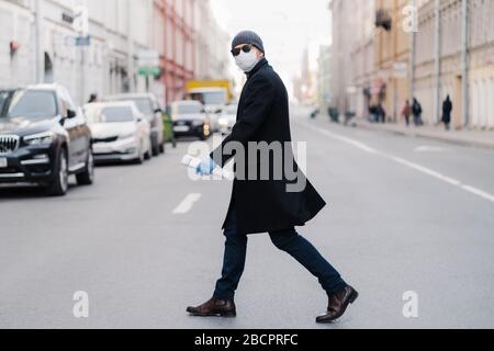 Virus covid -19. L'uomo attraversa la strada, vestito con cappotto nero, cappello e occhiali da sole, indossa la maschera totettiva per la protezione da virus pandemico, posa all'aperto a bu Foto Stock
