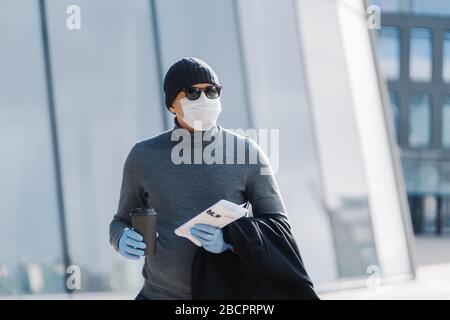 Colpo orizzontale di uomo caucasico vestito con dolcevita e cappello, toglie il cappotto, indossa maschera medica protettiva e guanti di gomma durante la situazione epidemica Foto Stock