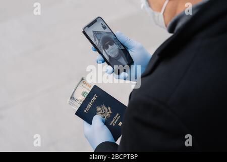 Scatto di uomo irriconoscibile seguire le regole di distanza sociale, fa videochiamata, tiene il telefono cellulare, passaporto con soldi indossa guanti protettivi passeggiate Foto Stock