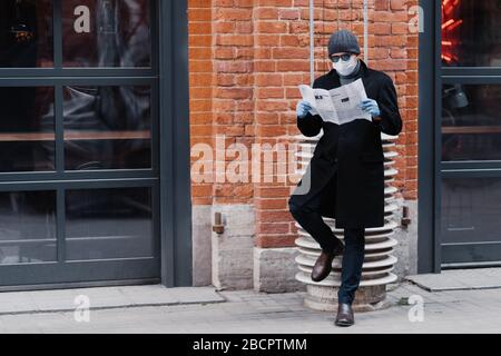 Scatto a lunghezza intera di uomo serio vestito in cappotto nero, indossa occhiali da sole e maschera medica, legge giornale, posa contro muro di mattoni, si previene Foto Stock