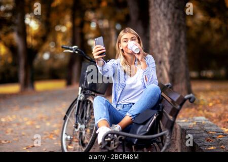 Ragazza giovane bionda con caffè a portata di mano prendendo selfie dal telefono cellulare in parte con bicicletta sullo sfondo Foto Stock