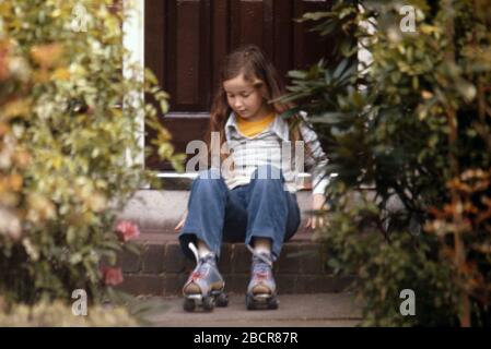 Una ragazza di 9 anni con i capelli scuri lunghi che portano i jeans blu del denim nel giardino anteriore della sua casa della famiglia, seduta sul gradino appena circa per ottenere in su sui suoi pattini del rullo 1974 Regno Unito Foto Stock