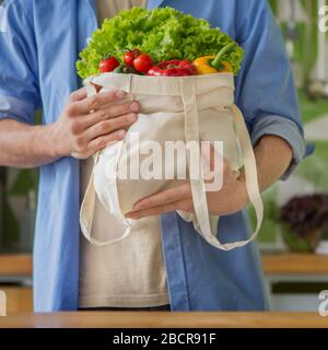 Uomo che tiene grande sacchetto riutilizzabile di cotone pieno di verdure in cucina Foto Stock
