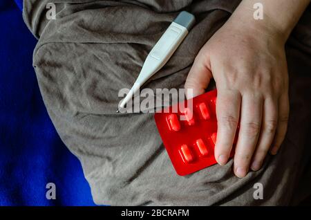 Blister con pillole in una mano femminile. La palma femminile poggia su un blister con pillole. Il termometro è nelle vicinanze. Vista dall'alto ad angolo. Messa a fuoco selettiva. Foto Stock