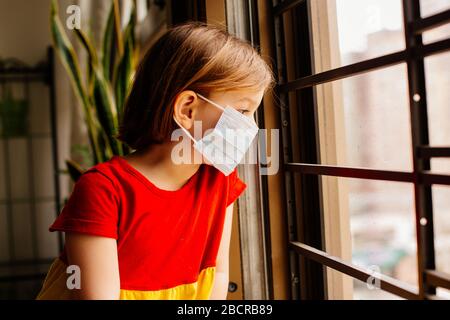 Bambino piccolo con protezione antivirus maschera chirurgica del viso che guarda fuori la finestra, rimanendo a casa per le distanze sociali a causa di coronavirus Foto Stock