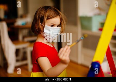 Ritratto di un piccolo bambino prescolare con protezione antivirus maschera facciale chirurgica a causa di coronavirus, pittura in un cavalletto artistico a casa Foto Stock