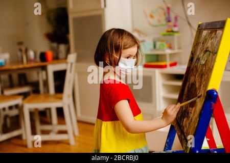 Ritratto di un piccolo bambino prescolare con protezione antivirus maschera facciale chirurgica a causa di coronavirus, pittura in un cavalletto artistico a casa Foto Stock