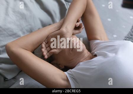 Donna anziana che copre gli occhi con le braccia, cercando di dormire in camera da letto. Foto Stock