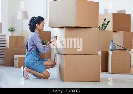 Vista laterale ritratto a tutta lunghezza di giovane donna asiatica che scrive su scatole di cartone che li etichettano per muoversi fuori, copia spazio Foto Stock