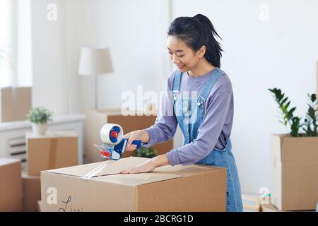 Ritratto di donna asiatica sorridente imballaggio scatole di cartone con dispenser di nastro mentre st si sposta verso casa nuova, copia spazio Foto Stock