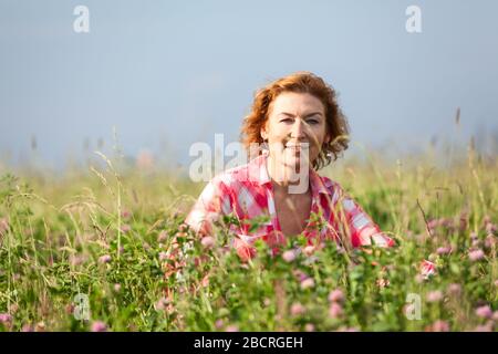 Donna matura vestita camicia rossa seduta in erba profonda e verde sul prato. Femmina caucasica, guardando la macchina fotografica Foto Stock