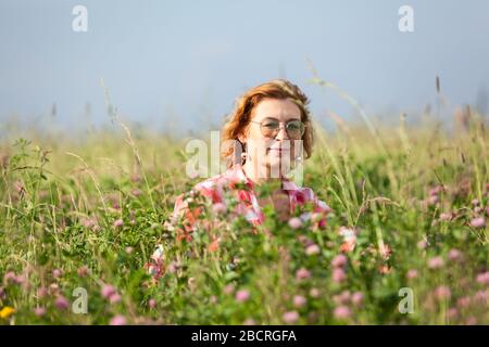 Donna caucasica matura in camicia rossa e occhiali seduti in erba alta sul campo, guardando la macchina fotografica Foto Stock