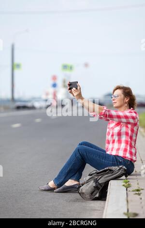 Donna attraente blogging con smartphone mentre si siede al confine della strada in città, autostrada vuota Foto Stock