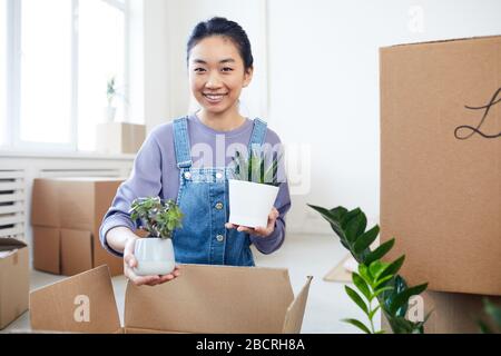 Ritratto di giovane donna asiatica imballaggio piante a scatole di cartone e sorridente alla macchina fotografica mentre si sposta a casa nuova o appartamento, copia spazio Foto Stock