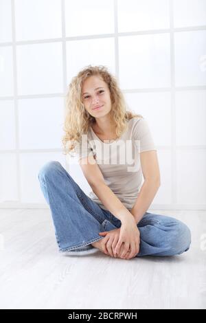 ritratto di giovane donna seduta sul pavimento vestita casual con capelli rossi ricci e lunghi isolati su sfondo bianco finestra Foto Stock