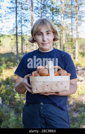 Donna di mezza età che tiene il cesto pieno di funghi con tappi di arancia, si trova nella foresta Foto Stock