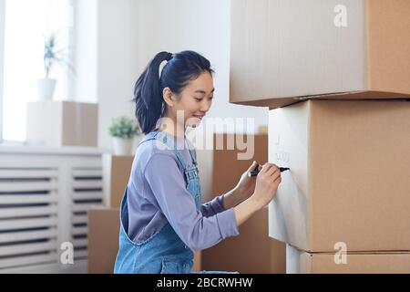 Ritratto di vista laterale di donna asiatica sorridente che scrive sulle scatole di cartone che li etichettano per muoversi verso la nuova casa, spazio della copia Foto Stock