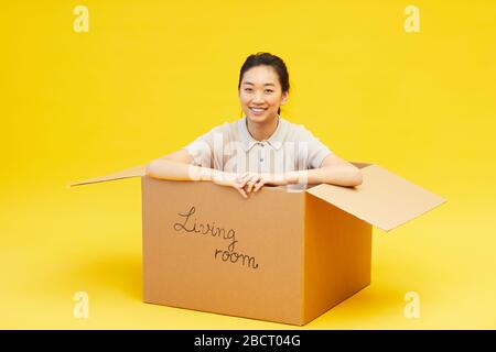 Ritratto a tutta lunghezza di giovane donna asiatica seduta in scatola e sorridente alla macchina fotografica, studio girato su sfondo giallo luminoso, copia spazio Foto Stock