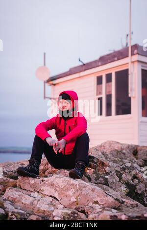 Donna seduta sulle rocce guardando il tramonto sulla baia di St. Brides alla stazione meteorologica di Martin's Haven - Pembrokeshire Coast Path, West Wales UK. Foto Stock