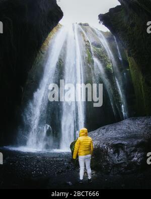 Ragazza in piedi sotto una grande cascata in una giacca gialla. Enorme cascata di Gljufrabui, situata accanto alla cascata Seljalandsfoss, famosa per i turisti Foto Stock